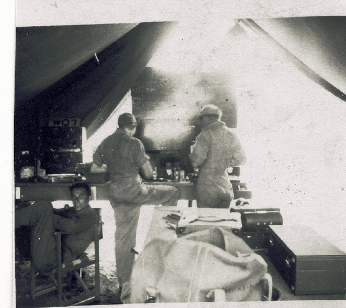 Three men in a tent fitted out as a radio shop: two work standing at a bench of receivers and test gear, while a third sits back in a folding canvas chair and looks at the camera. A small plate on one set is lettered "WO7", and a chalked board hangs on the tent wall behind the bench.