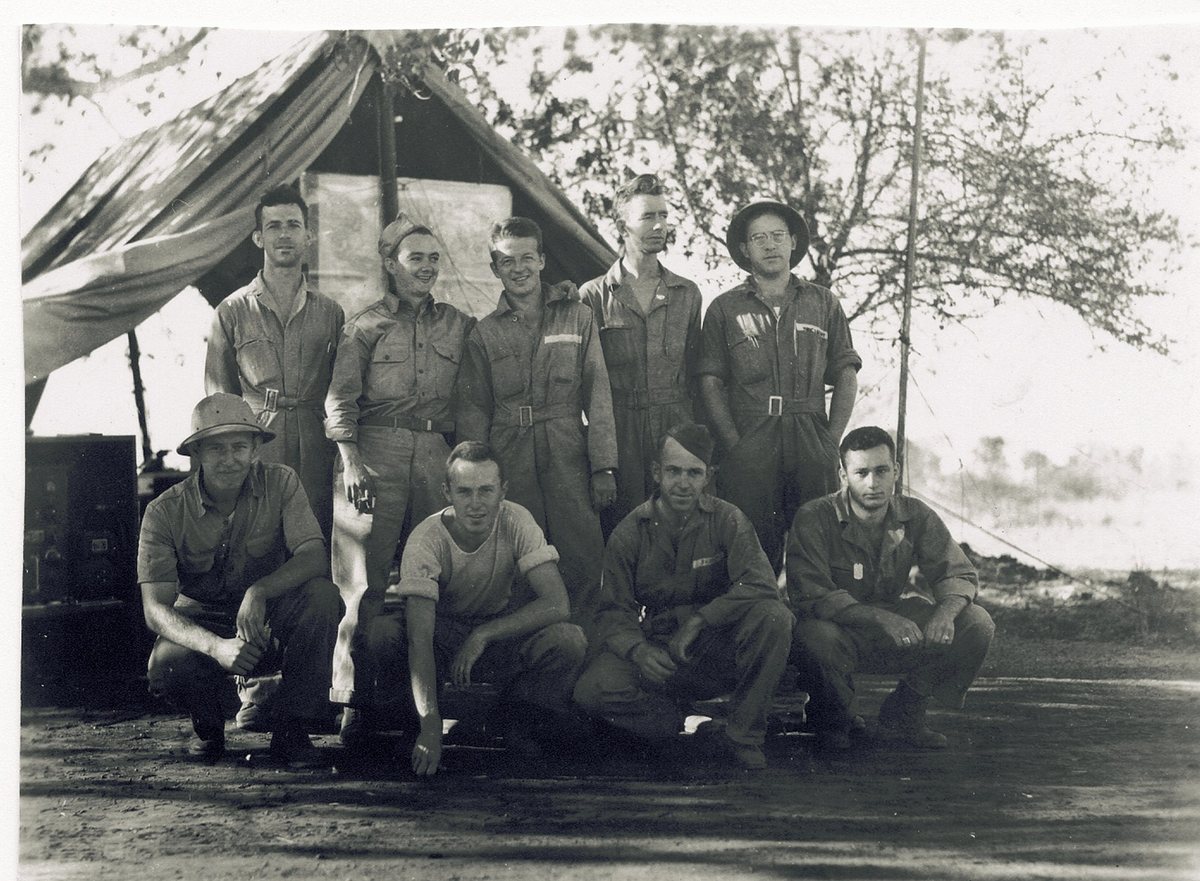 Nine men, five standing and four crouching, in front of a tent with its side flap propped open on a pole and a large pale chart pinned up inside. A dark equipment case fitted with dials and small panels stands on the ground at the left, and a slender antenna mast with its wire rises at the right of the group; two of the men wear sun helmets and one of those crouching is in a T-shirt.