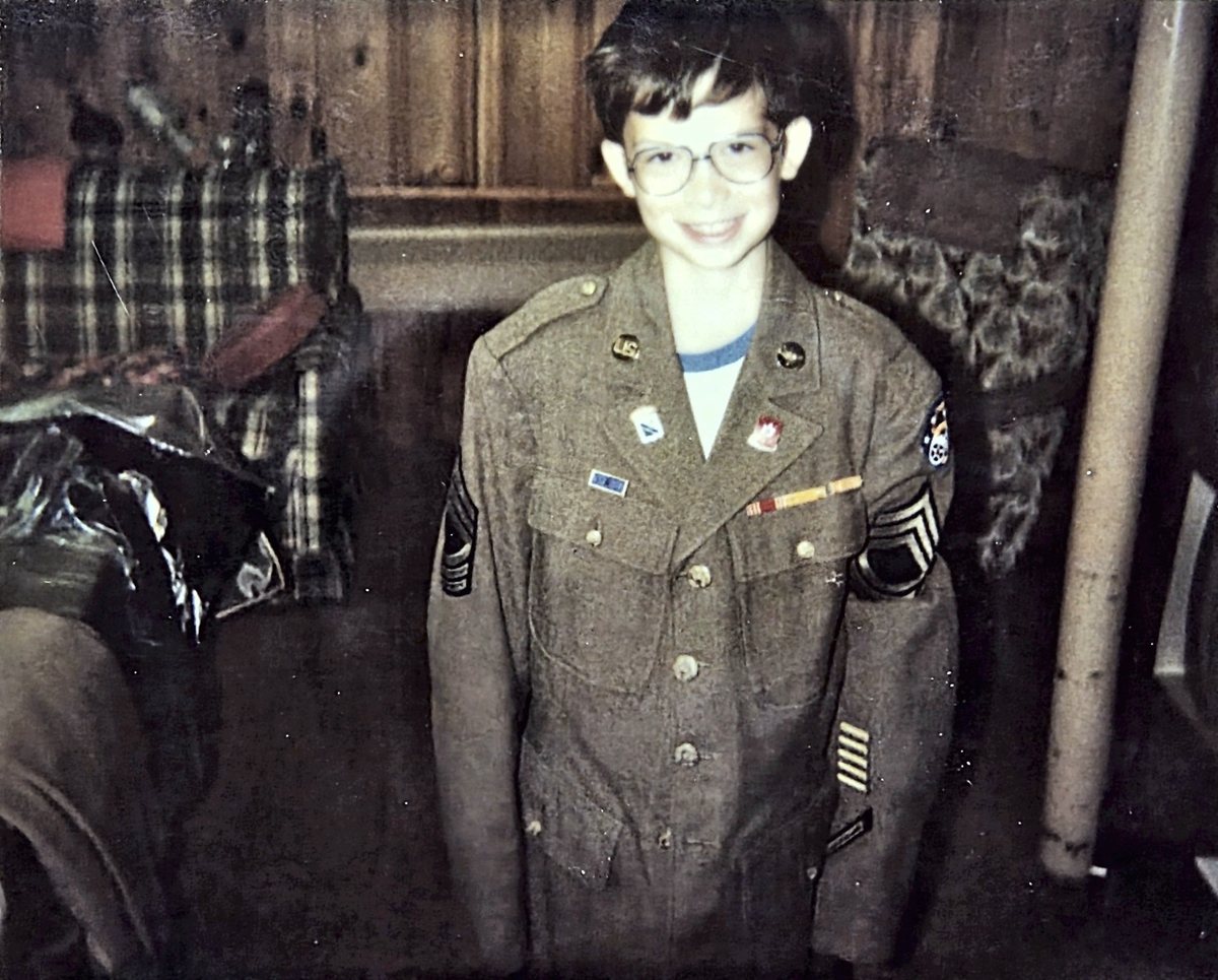 A boy in glasses stands in a panelled room wearing an oversized Army olive-drab service coat, its master sergeant chevrons on both sleeves, "U.S." and squadron-style insignia discs on the lapels, a ribbon bar and a shoulder patch on the left arm; the back identifies him as Allen's grandson Jeremy, in 1986, wearing Allen's own coat while going through the war collection.