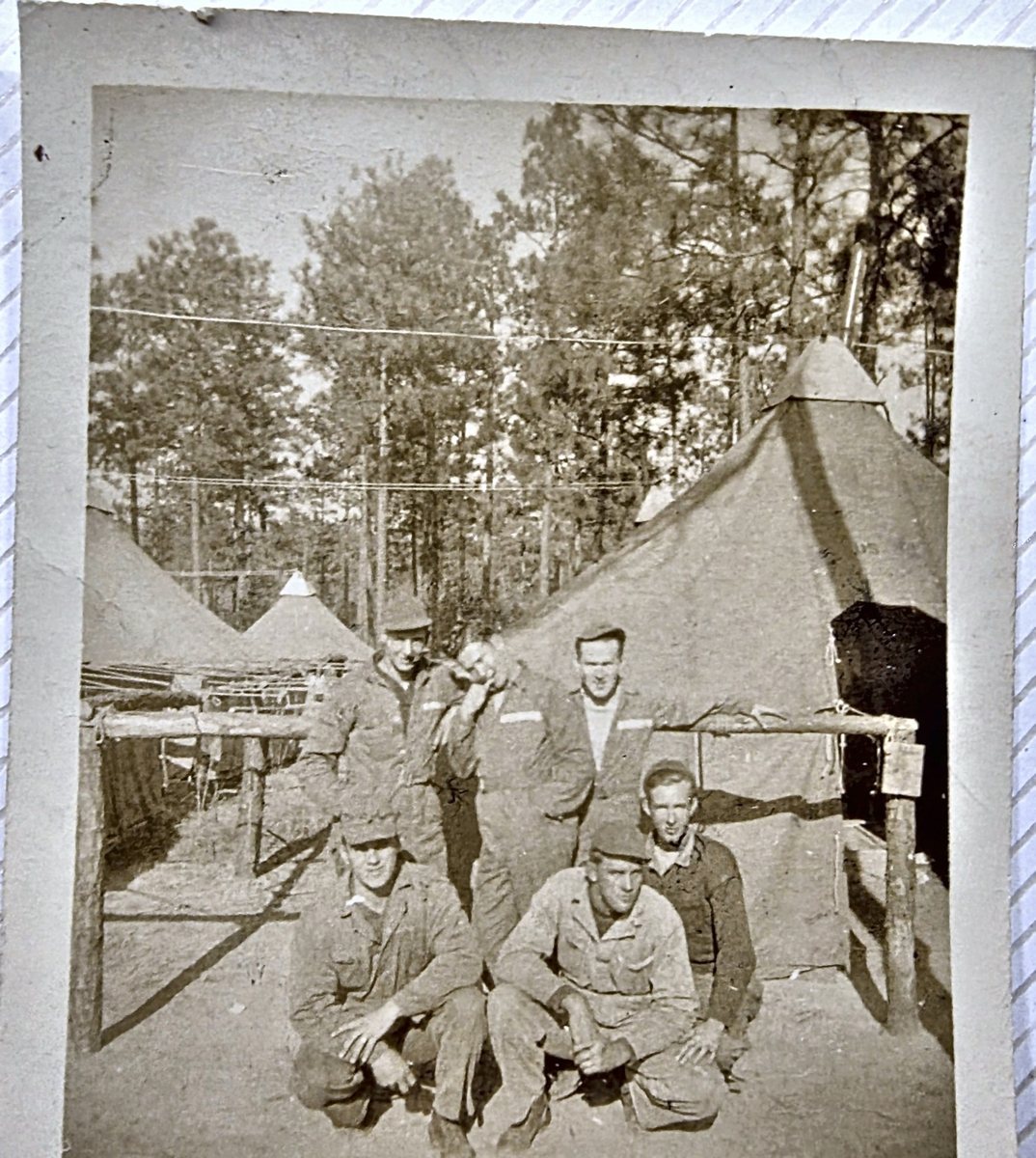 Six men pose among pyramidal tents pitched in a pine wood, four standing -- one of them stooped forward with his head down -- and two crouching in front. Five wear work fatigues and soft caps; the man at the right is bareheaded in a dark sweater with a light scarf at his neck. A rough timber rack and table stand at the left, a timber rail runs across the group, and wires are strung overhead. The back reads "Pope Field / Maneuvers / Nov. 1941".