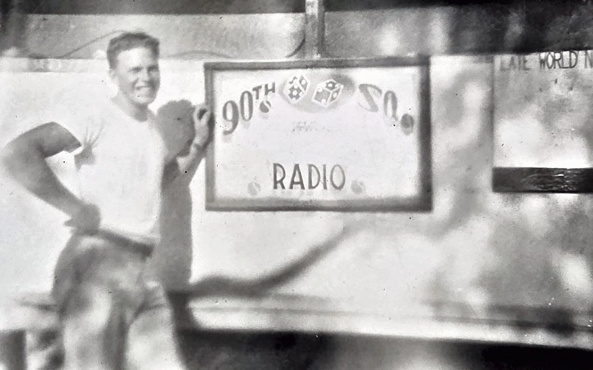 A grinning man in a white T-shirt, hand on hip, stands beside a framed hand-painted sign reading "90TH SQ." with a pair of dice and, below, "RADIO"; a board at the right is lettered "LATE WORLD N...". The back carries a large flourished pencil signature -- read as "John B. H----" by one reader and as wholly illegible by the other -- plus part of a censor stamp, so the man is not named.