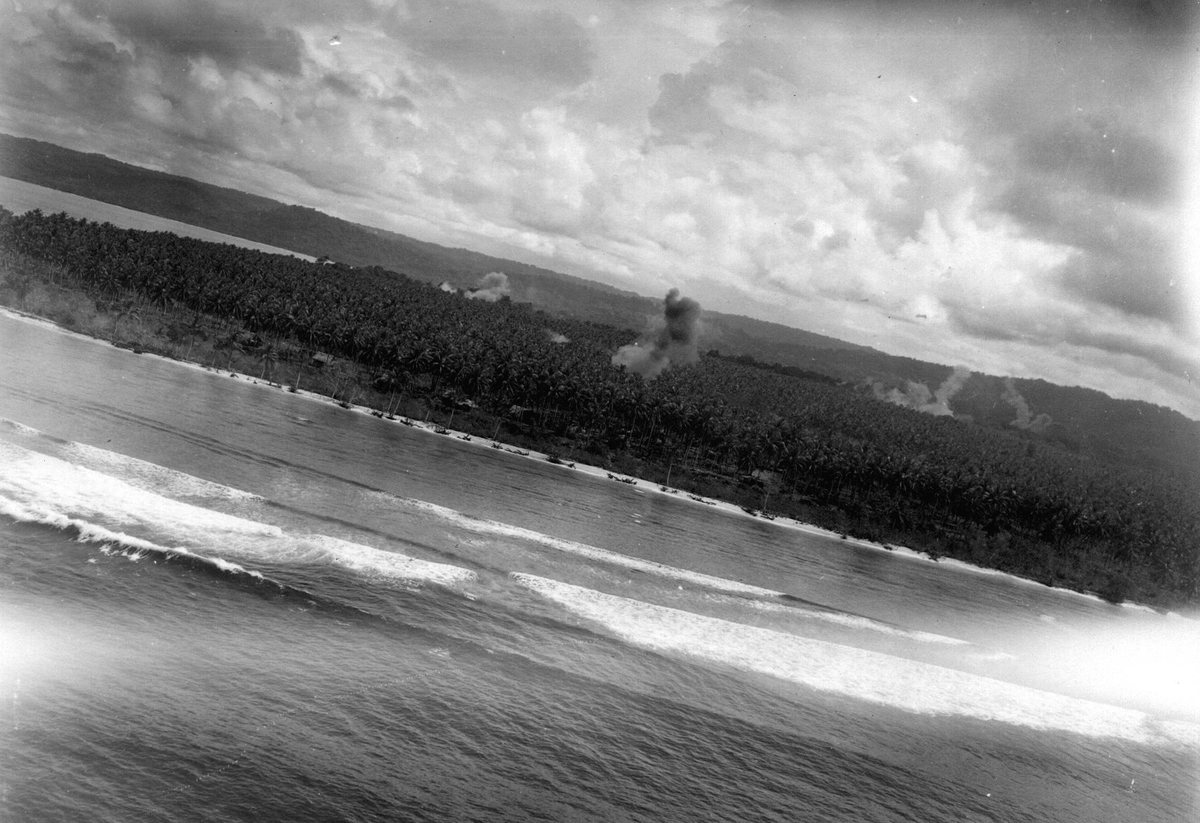 An attack in progress on a New Guinea beachhead, photographed from the air — smoke rising from the tree line as landing craft sit offshore.