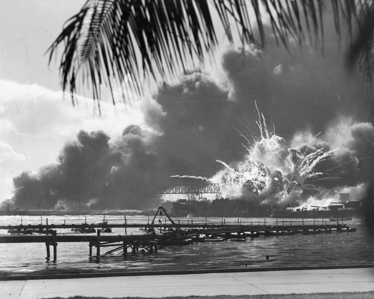 A harbour under attack, photographed from the shore beneath a palm frond: a white fireball and streaks of burning debris burst from a warship inside a floating drydock, heavy black smoke rolling across the water behind a pipeline trestle. Identified in the U.S. Navy record (National Archives 80-G-16871) as the destroyer USS Shaw in floating drydock YFD-2, Pearl Harbor, 7 December 1941.