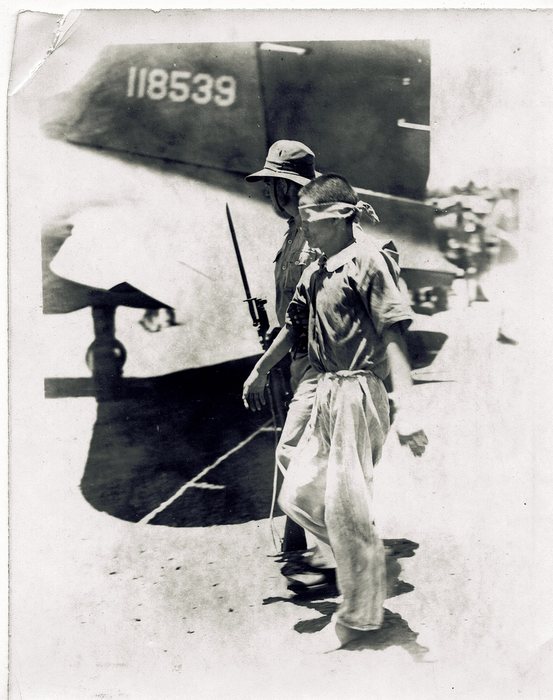 Armed soldiers escort a Japanese prisoner of war past an aircraft.