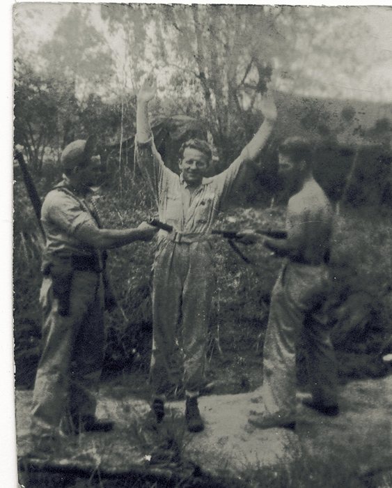 Three men pose for the camera in a scrubby clearing: the man in the centre stands grinning with both hands raised, while the two flanking him level rifles at him, one in shirt and cap with a second rifle slung on his shoulder, the other stripped to the waist.