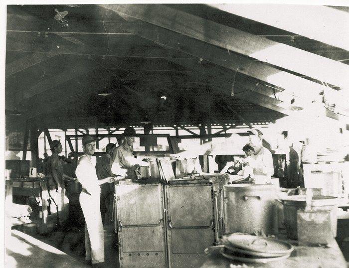 A field kitchen under a timber-truss roof open along both sides, with cooks in white aprons and men in fatigues working over field ranges; large aluminium stock pots, open cans and stacked pan lids crowd the benches in the foreground.