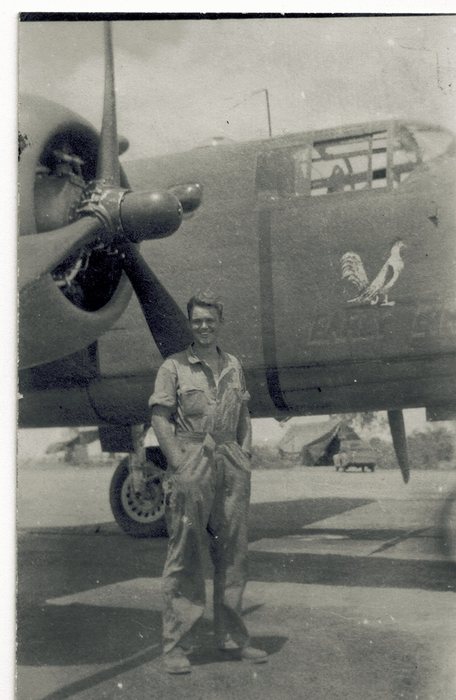A serviceman in oil-stained coveralls stands between the propeller and the nose of a combat aircraft, with a white rooster painted on the nose above lettering now too faded to read. A tent and a truck stand at the edge of the strip behind him.
