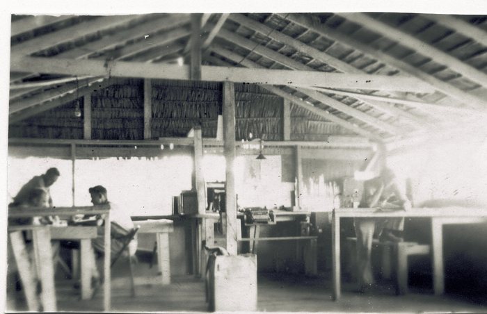 Three men at work inside an open-sided building with a thatched roof on rough timber posts: two at a table on the left, one of them seated in a canvas chair, a typewriter on a bench in the middle of the room, and a third standing at a bench on the right. The glare through the open walls swallows the far end, so it cannot be said whether others are in the room.