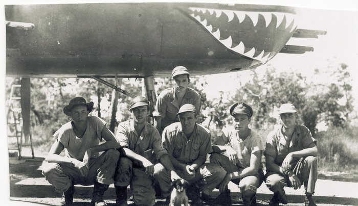 Five men crouch in a row beneath the nose of an aircraft while a sixth stands behind them, the nose painted with a shark's mouth of white teeth and four machine-gun muzzles projecting past it. A small dog sits at the feet of the two men in the centre.