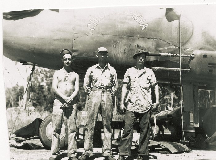 Three men stand under the nose of a bomber lettered in outlined capitals, the first letters reading CHIC and the last, past a worn stretch, HIYA; the man nearest the camera is stripped to the waist and wearing dog tags. Two pairs of machine-gun barrels project from the gun pack on the fuselage side, with the main wheel and a tarpaulin behind them.