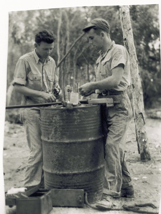 Two men work over an upturned oil drum standing in for a bench, with a vise clamped to a board across the top of it beside an oil can and a small bottle; a metal tool box sits on the ground and a length of rod rests across the drum.