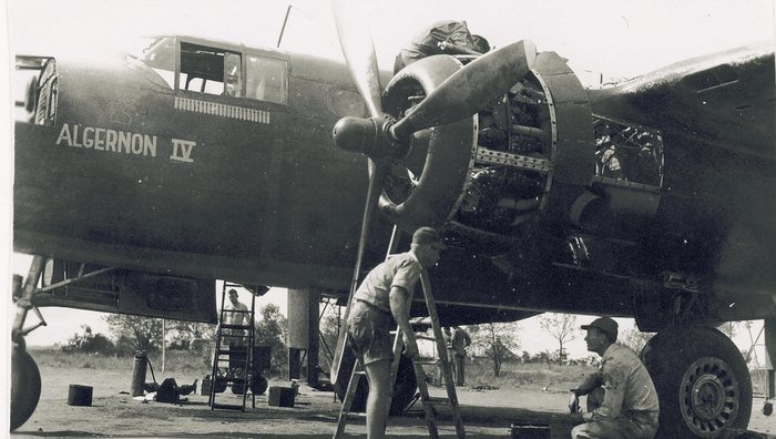 Engine maintenance on a bomber lettered <b>ALGERNON IV</b> in block capitals with the Roman numeral. A faint outline cartoon figure is sketched on the skin above and left of the name, and a row of mission symbols runs under the cockpit.