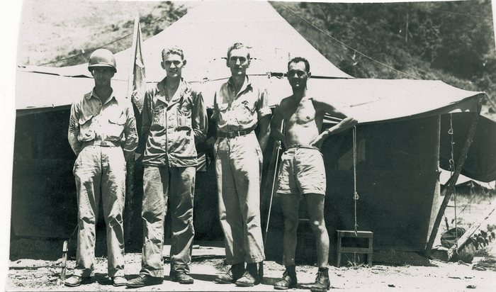 Four men stand in front of a squad tent pitched against a wooded hillside: one in a steel helmet with sergeant's chevrons on his sleeve, one in coveralls, one in shirt and trousers, and one bare-chested in shorts. A furled flag on a staff stands between the first two men, and a thin pole leans upright between the last two.