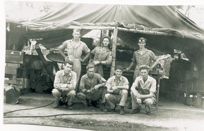 Seven servicemen at a maintenance area stocked with fuel drums.