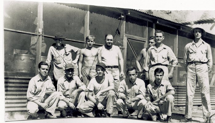 Eleven men pose outside a corrugated-iron hut with an open upper wall, five kneeling in front and six standing behind, in fatigue shirts, caps and bush hats, two of them stripped to the waist. A large urn stands on a bench at the left.