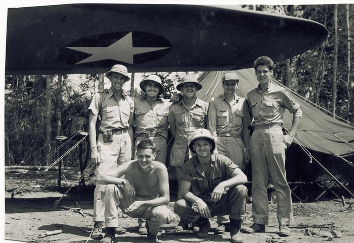 Seven servicemen grouped under the wing of a parked aircraft, the white national star insignia on the underside directly above their heads. Five stand in khaki shirts, bush hats and one sun helmet, while two crouch in front, one of them stripped to the waist; a squad tent and folding cots stand behind them at the edge of tall timber.