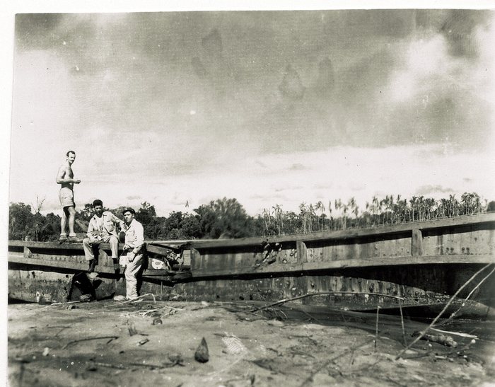 Three servicemen on a large wreck of heavy timber and riveted steel plate, snapped through near its centre and lying on a flat of dried mud and sand. One stands on top stripped to the waist in shorts, one sits on the broken edge in fatigues, and one stands below it wearing a cap; a line of palms runs along the horizon.