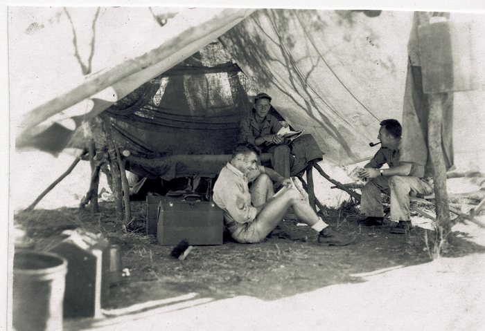 Three servicemen relax inside a tent, one smoking a pipe.