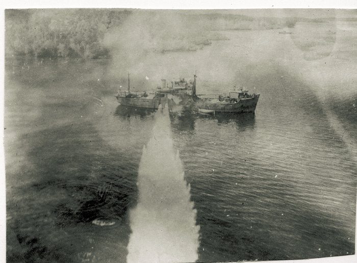 A low oblique aerial photograph of two small coastal vessels lying close alongside one another in sheltered water, with smoke or steam rising between their hulls, a boat made fast on the outboard side, and a narrow straight track of white water running across the surface toward them. A mangrove shoreline fills the top of the frame.