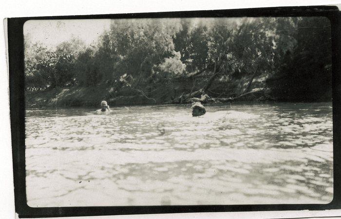 Three men at a bathing place on a wide, slow river: two out in the water with only their heads and shoulders showing, and a third sitting bare-chested on a fallen tree that leans out over the surface from the scrub of the far bank.