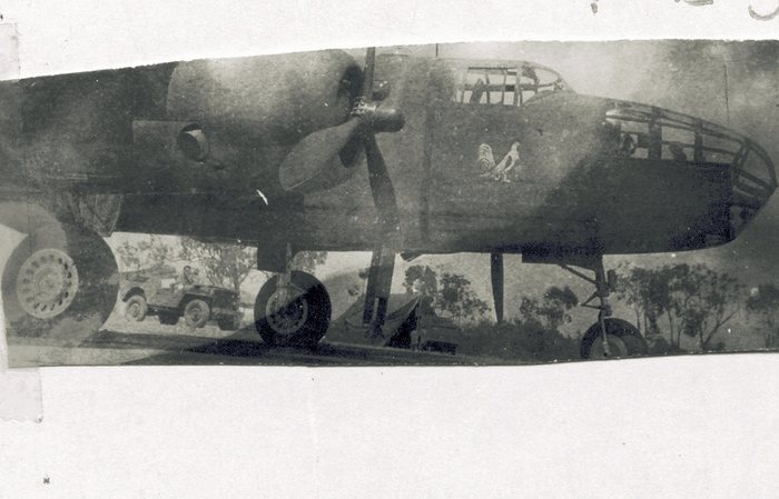 A twin-engined bomber standing on a dispersal, photographed from beneath the wing, with a white cockerel painted on the nose ahead of the cockpit glazing; a jeep with a driver at the wheel is parked under the fuselage, and scrub and gum trees stand beyond.