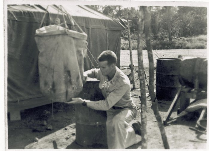 A serviceman in light one-piece coveralls kneels at the end of a tent beside a field wash line, one hand steadying a board across a galvanised can while a canvas tub hangs above it from a rope bridle; oil drums and a shallow pan stand on the bare ground behind.