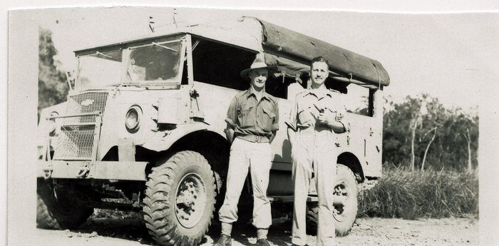 Two servicemen in khaki stand beside a snub-nosed military lorry with a wire-mesh grille guard, a canvas cab top and heavy off-road tyres, on a stony clearing at the edge of dry scrub; the man on the left wears a broad-brimmed field hat, the man on the right holds something small in both hands.