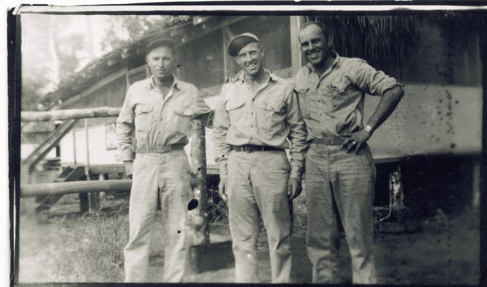 Allen at the right of three servicemen in khaki shirts and trousers, two of them in field caps, standing shoulder to shoulder in front of a timber building raised on posts with part of its roof thatched; a log rail and steps run up to the building on the left.