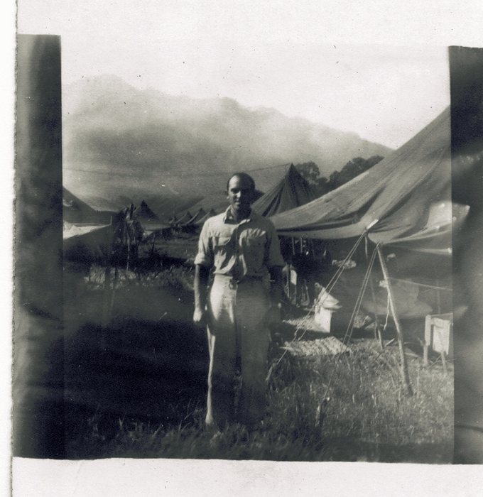 Allen, bareheaded, in a light shirt and trousers in a camp of pyramidal and tarpaulin tents, with a ridge of cloud-covered high ground behind him. Guy ropes run down at right, where a pale bag and a flat woven mat lie on the ground under the tent fly.