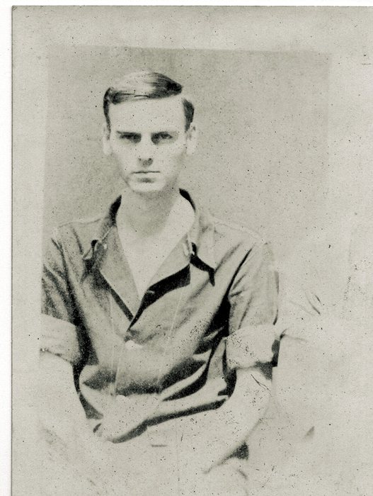 A waist-up portrait of a young serviceman, his hair combed back, seated in an open-collar shirt with the sleeves rolled to the elbow, his forearms crossed and hands clasped in his lap.