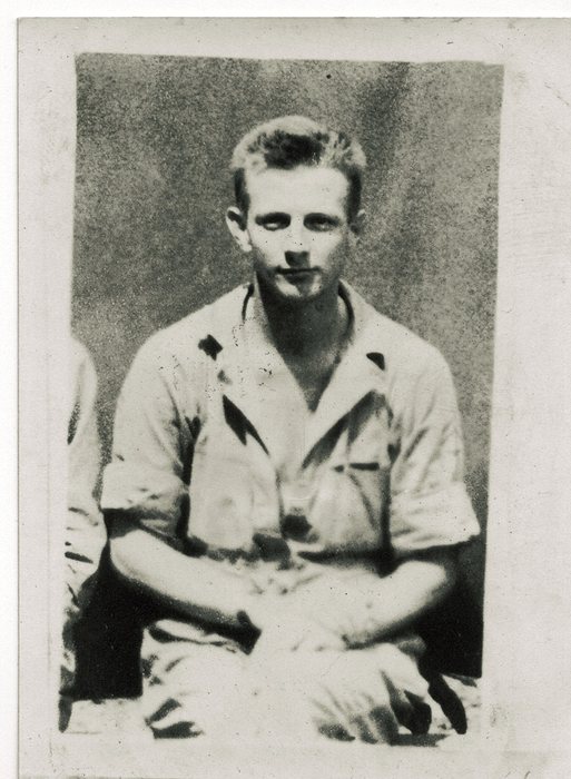 A young serviceman sits with his arms crossed over his knee in a light open-collar shirt with the sleeves turned up; a second man's arm and shoulder are cut off at the left edge of the frame.