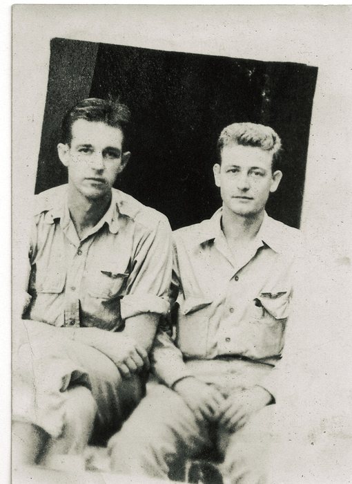 Two young servicemen sit side by side in front of a dark doorway, both in light open-collar summer shirts with the sleeves rolled. A small winged badge is pinned above the left man's breast pocket; the print is too coarse to tell which qualification wings they are.