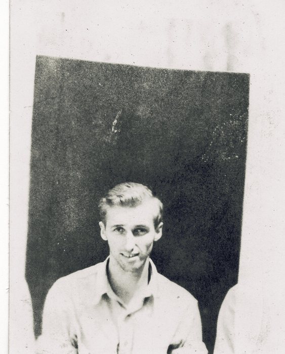 A young serviceman in a light open-collar shirt sits smiling against a dark panel or wall; the shoulder of another man is just visible at the right edge.
