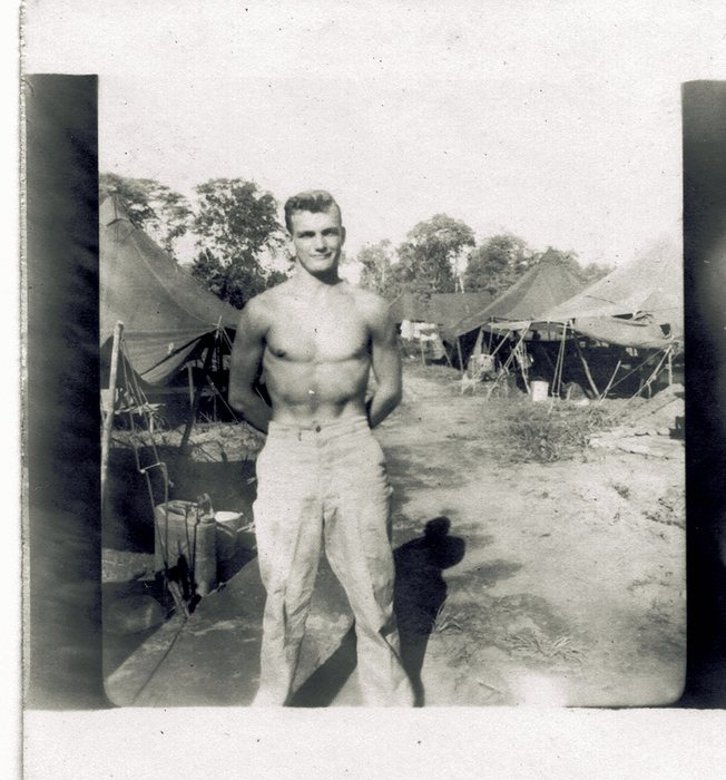 A shirtless serviceman relaxes in camp, tents visible behind him.