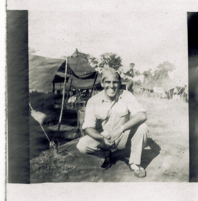 Allen in a garrison cap, light shirt and trousers, crouching on bare ground in front of a large tarpaulin shelter rigged on poles, with further tents and eucalyptus-like trees beyond.