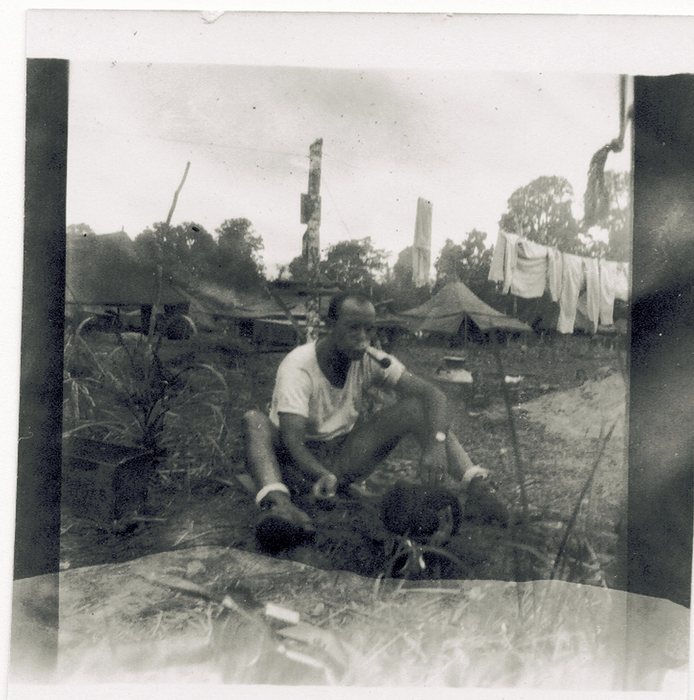 Allen sitting on the ground in a white undershirt with a pipe in his mouth and a towel round his neck, wearing boots with white socks turned down over the tops, in a camp of thatched and canvas shelters. A laundry line of shirts and towels is strung at the right, and behind him two tall stripped poles carry thin wires, one with a small box fixed part way up.