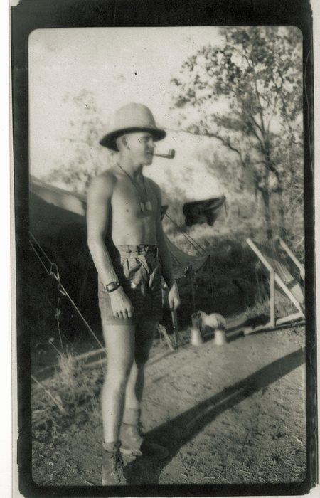A serviceman stands stripped to the waist in a pith helmet with a pipe in his teeth, wearing cut-off shorts, a wristwatch and boots, with identity tags on a chain around his neck. Behind him are a tent, mosquito netting and a canvas deck chair, and two pale tins stand on the bare ground at his feet.
