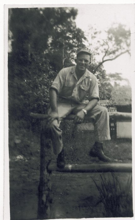 Allen in a peaked fatigue cap and work shirt, sitting on a rough timber rail with one boot up, dense vines and jungle foliage behind him and a low table or crate at right.