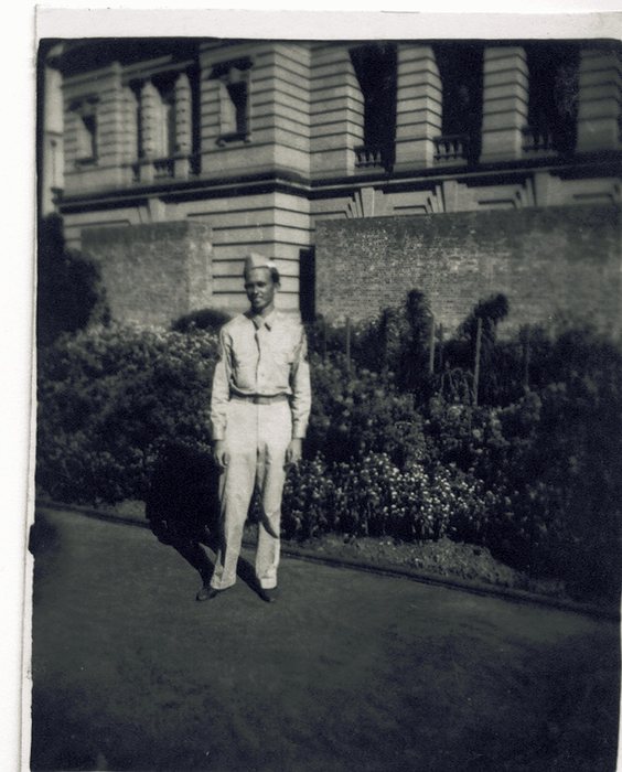 A serviceman in light summer khakis and a garrison cap stands on a paved garden path, a low brick wall and dense flowering shrubs behind him. Above the wall rises the rusticated masonry facade of a large building, its tall windows fronted by small stone balustrades, the top of it cut off by the frame.