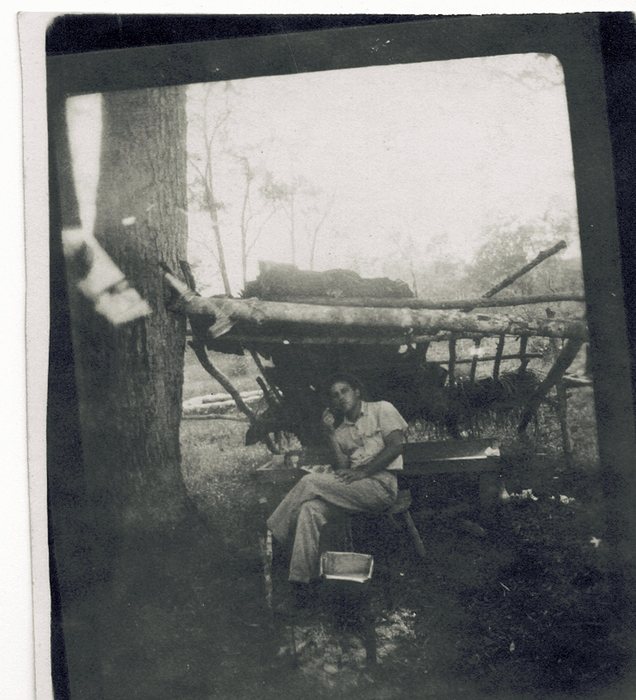 Allen in shirt and trousers on a folding chair beneath a shelter built of poles and split logs lashed against a large tree, papers spread on the bench beside him and a white enamel pan on the ground at his feet.