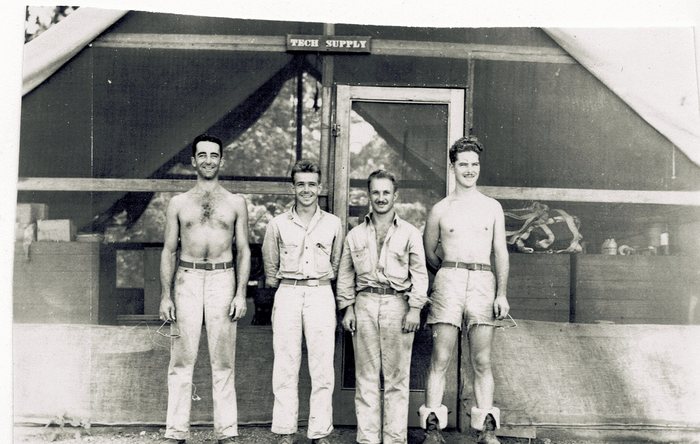 Four servicemen stand in a row in front of a screened tent-front hut, beneath a small painted board over the doorway reading "TECH SUPPLY". Two are bare-chested, one of them in cut-down shorts with boots and rolled-down socks, and a parachute harness, cartons and cans sit on the shelving behind them.
