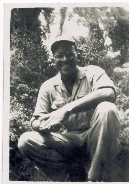 Allen, in spectacles and a soft fatigue cap, sitting on the ground against a wall of jungle growth, sleeves pushed above the elbow and his wristwatch on his left wrist. Identified by the family.