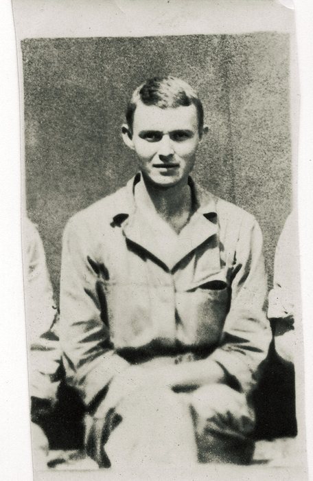 A young serviceman in a light shirt and trousers, seated with his hands on his knees against a plain stucco wall. The shoulders of the men on either side of him show that this small print was trimmed out of a larger group photograph.