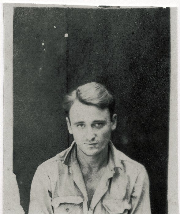 Close portrait of a fair-haired serviceman in an open-collar shirt, seated in front of a dark wall; a neighbour's shoulder at the lower left corner shows the print was cut from a group photograph.