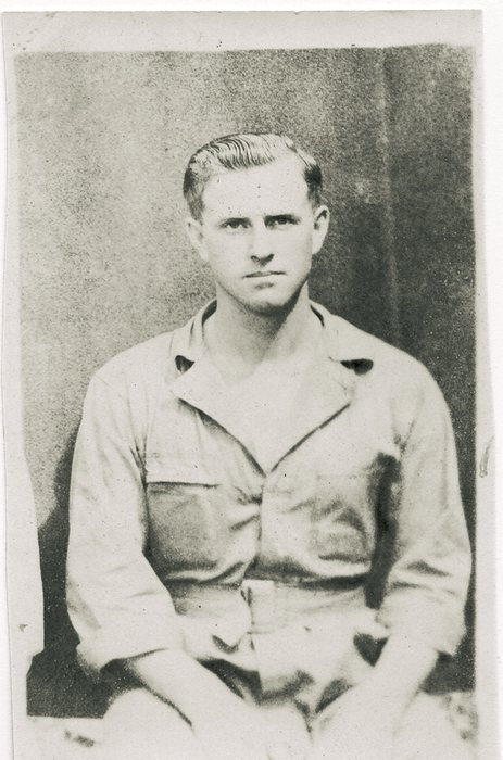 A serviceman with his hair combed to one side sits with his hands in his lap against a mottled concrete wall, in the same light shirt and trousers worn by the other men in this set of trimmed cut-outs.