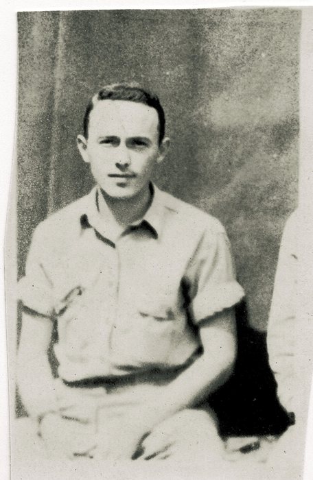 A slightly built serviceman sits in a short-sleeved shirt with the cuffs turned back and his hands in his lap, against a mottled wall; the arm and leg of the man beside him remain at the right edge of the trimmed print.