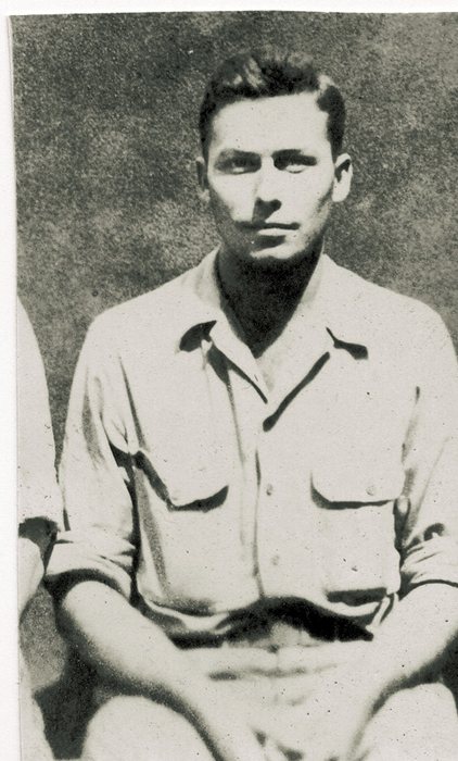 A serviceman in a two-pocket shirt with the sleeves pushed up sits with his hands in his lap in front of a grainy wall; the arm and knee of the man next to him are still visible at the left edge where the print was cut down.
