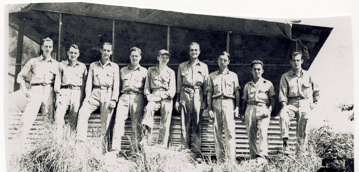 Nine men in khaki shirts and trousers along the front of a long camp building, its raised floor skirted with corrugated iron and its upper wall left as open framing, with tall grass growing up in front of them. One man, in a soft fatigue cap, sits perched on the sill with his legs crossed and his hands in his lap; several of the others wear spectacles.