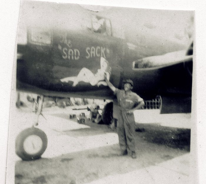 A serviceman in coveralls and a soft cap stands with one hand raised against the nose of a bomber lettered "SAD SACK" in quotation marks, with the cursive word "The" above it and a reclining pin-up painted below. A spare tyre stands upright on the ground at the left, and crates and equipment are stacked along the hardstand behind him.