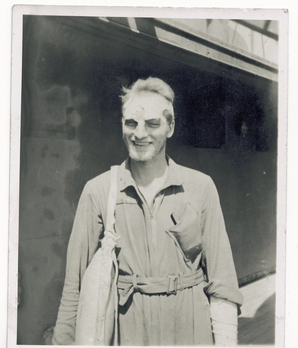 A serviceman in a belted one-piece work suit with a canvas bag slung across his shoulder, standing in front of a dark building opening, his hair, face and eyebrows caked with a pale dust or powder.